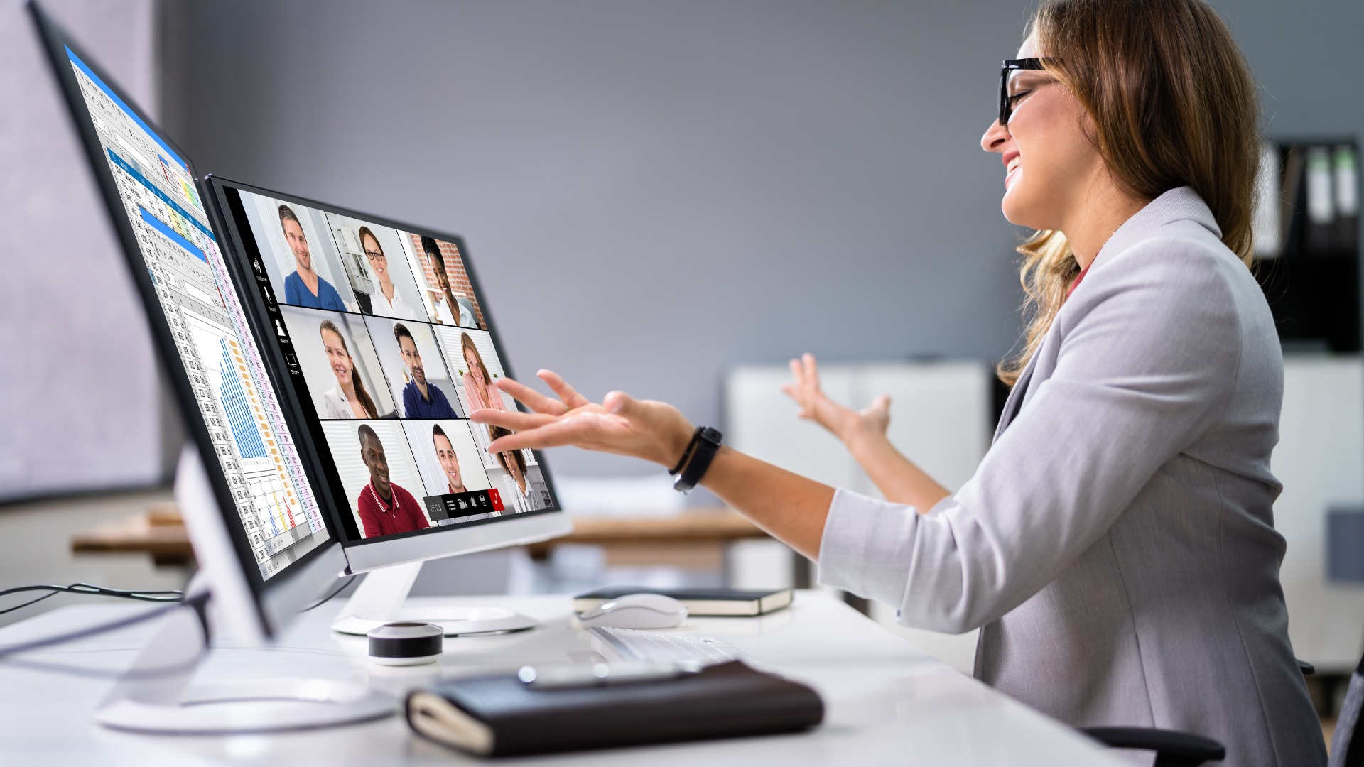 A professional woman gestures while engaging in a virtual meeting, with a monitor showing multiple participants in a video call — representing expert-led, collaborative online learning.